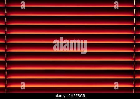 Window blinds are orange with a smooth transition to ruby red and black with ropes around the edges close-up Stock Photo