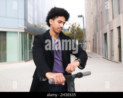 Stylish young African American male with curly dark hair checking time on smartwatch while riding electric scooter in city downtown Stock Photo