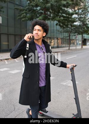 Full body of confident young African American male millennial with dark curly hair in stylish outfit talking on smartphone while standing on city stre Stock Photo
