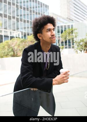 Serious young African American male entrepreneur with dark curly hair in stylish clothes leaning on glass table and looking away thoughtfully while dr Stock Photo