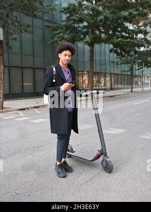 Full body of confident young African American male millennial with dark curly hair in stylish outfit messaging on smartphone while standing on city st Stock Photo