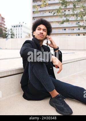 Young black man with dark Afro hair in trendy outfit talking smartphone on city square Stock Photo