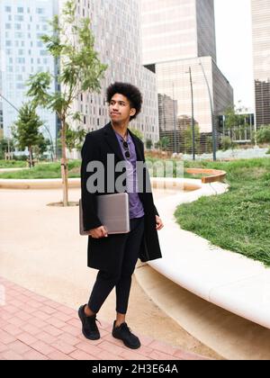 Full body of trendy young ethnic male freelancer with curly dark hair in stylish outfit walking in city park near contemporary skyscrapers with laptop Stock Photo