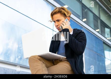 Focused young male freelancer in casual wear having phone conversation while sitting near modern building with netbook on street of city Stock Photo
