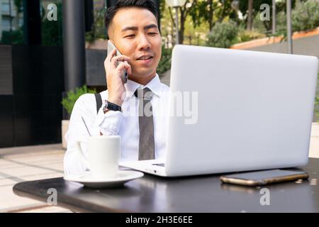 Young ethnic male entrepreneur with netbook talking on cellphone while sitting at urban cafeteria table with cup of coffee Stock Photo