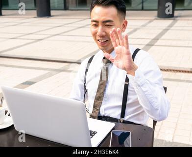 Content young Asian male entrepreneur waving hand during video chat on netbook at table in street cafe Stock Photo