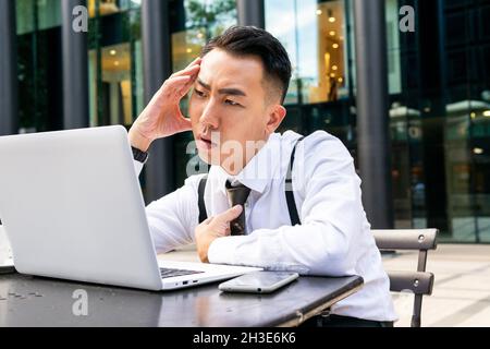 Thoughtful ethnic businessman in white shirt touching temple while sitting at table with netbook and smartphone in urban cafe Stock Photo