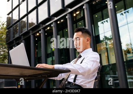 Young ethnic male entrepreneur surfing internet on laptop while sitting at urban cafeteria table Stock Photo