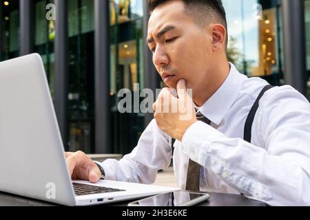 Crop pensive ethnic male entrepreneur in white shirt touching chin while working on netbook at cafeteria table in town Stock Photo