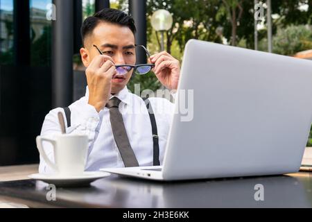 Well dressed young ethnic male entrepreneur putting on eyeglasses against table with netbook and hot drink in street cafeteria Stock Photo