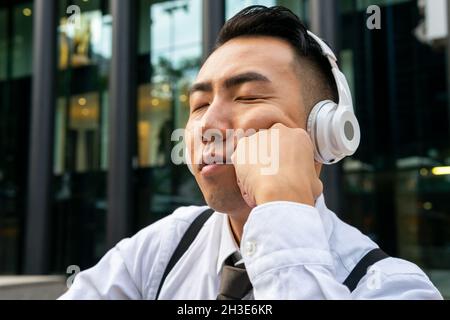 Tired young Asian male entrepreneur with closed eyes enjoying song from wireless headphones against city street Stock Photo