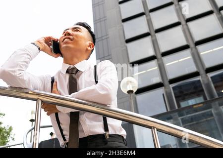 From below of contemplative young ethnic male entrepreneur leaning on fence while speaking on cellphone and looking away in city Stock Photo