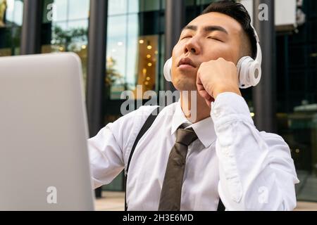 Mindful young Asian male entrepreneur with closed eyes enjoying song from wireless headphones against laptop in street cafeteria Stock Photo