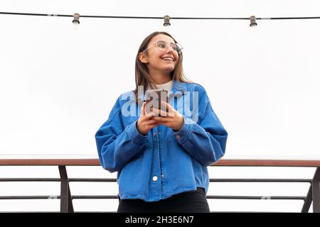 Smiling young female in casual outfit and eyeglasses standing in town street while surfing on cellphone under some hanging bulbs Stock Photo
