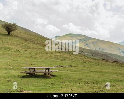 Hilly area covered with grass with wooden table and bench located against cloudy sky in countryside on summer day in nature Stock Photo