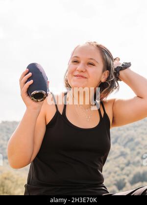 Positive female with bluetooth portable speaker looking away while sitting against forested mountain ridge with green trees trees in nature Stock Photo