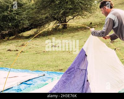 Side view of young male traveler in casual clothes opening camping tent on green grassy meadow during trip in countryside Stock Photo