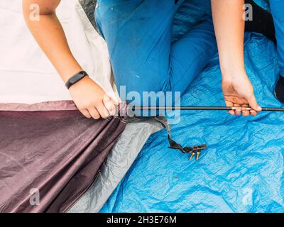 Crop unrecognizable female traveler in casual clothes and socks sitting on tent while assembling it after hiking on sunny day Stock Photo