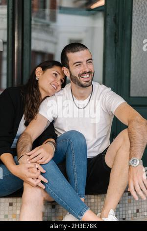 Content young ethnic couple with dark hair in casual clothes smiling and looking away while sitting together on stairs Stock Photo
