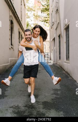 Full length of positive young Hispanic male millennial in casual clothes giving piggyback ride to cheerful laughing girlfriend on narrow street on sun Stock Photo