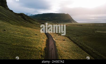 Breathtaking drone view of unrecognizable traveler walking along road near grassy hills during hiking trip in Iceland under cloudy sunset sky