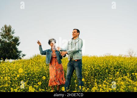 Happy young man in casual clothes opening bottle of champagne during romantic date with cheerful African American girlfriend in blooming meadow in cou Stock Photo