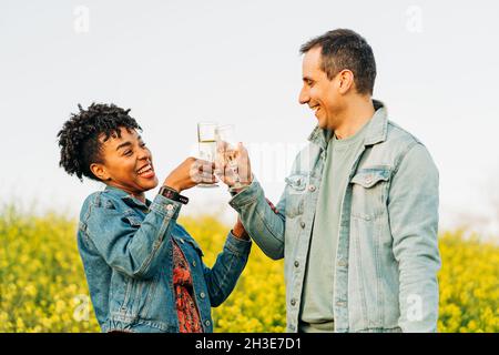 Cheerful young man and black Woman in denim jackets smiling and clinking glasses of champagne while standing in blooming green meadow during romantic Stock Photo