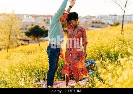 Happy young diverse couple holding hand while dancing together in blooming meadow during romantic picnic on sunny day Stock Photo