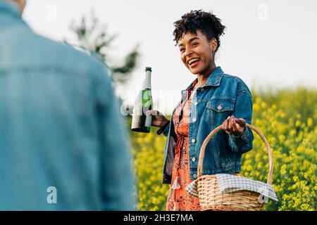 Cheerful young black female with curly hair in stylish dress and denim jacket smiling happily and looking at crop unrecognizable boyfriend while stand Stock Photo