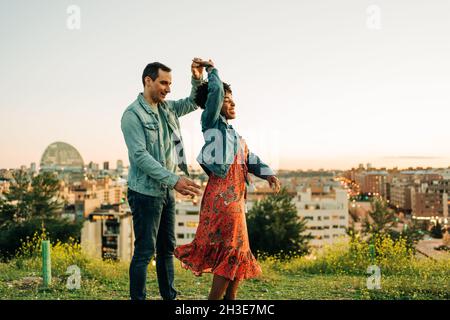 Happy young diverse couple holding hand while dancing together in blooming meadow on sunny day Stock Photo
