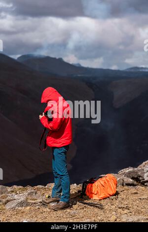 Side view of male photographer in outerwear standing on top of rocky cliff near active volcano Fagradalsfjall with black lava in Iceland in daytime Stock Photo