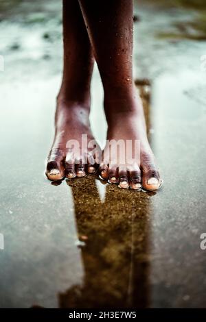 From above of crop unrecognizable black barefoot kid standing in small puddle on asphalt road on street on São Tomé and Príncipe island in daylight Stock Photo