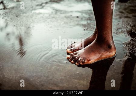 From above of crop unrecognizable black barefoot kid standing in small puddle on asphalt road on street on São Tomé and Príncipe island in daylight Stock Photo