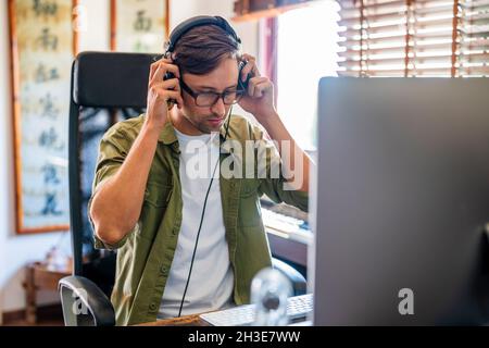 Male putting on headphones while sitting on chair at table and working on computer near window Stock Photo