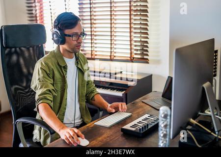 Side view of concentrated guy with headphones sitting at table and using mouse while working on computer Stock Photo