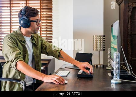 Side view of concentrated guy with headphones sitting at table and using mouse while working on computer Stock Photo