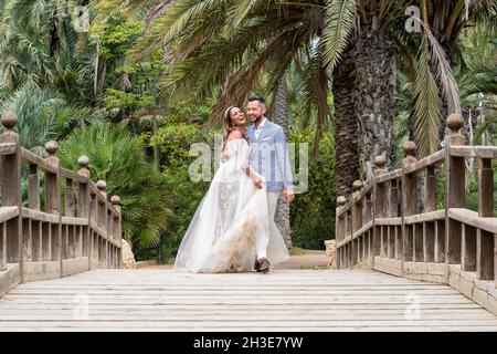 Married couple in wedding outfits standing on wooden footbridge with railing while embraced and looking away near green palms and plants in garden in Stock Photo