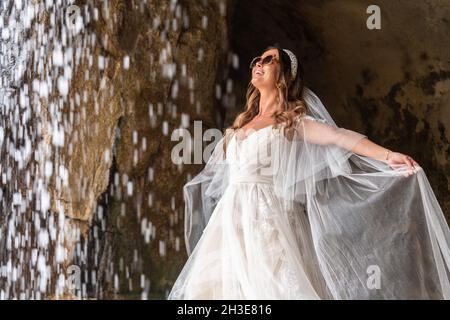 Bride in white wedding dress with veil and sunglasses standing on boulder  near falling cascade in nature during holiday celebration on summer day  Stock Photo