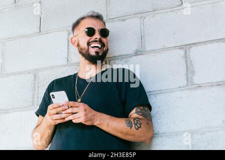 Happy bearded guy with tattoos in black t shirt and sunglasses standing near wall of building and using a smartphone in daylight Stock Photo