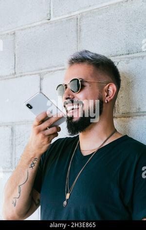 Happy bearded guy with tattoos in black t shirt and sunglasses standing near wall of building and recording audio message on smartphone in daylight Stock Photo
