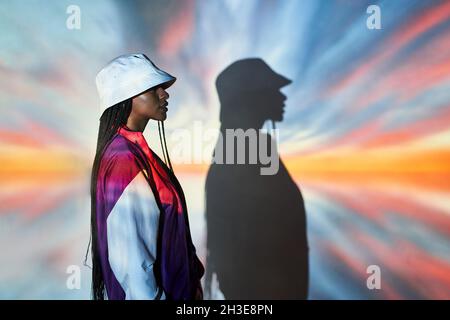 Side view of self assured young Dominican female teenager in trendy outfit and hat standing near wall with creative abstract projections and looking a Stock Photo