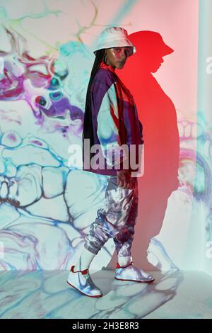 Side view of self assured young Dominican female teenager in trendy outfit and hat standing near white wall with creative abstract projections and loo Stock Photo