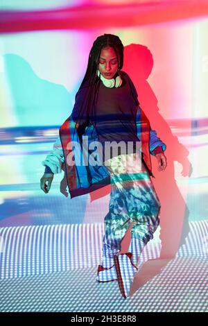 Full body of confident young ethnic female millennial in fashionable outfit and headphones standing on tiptoes near wall with colorful projection and Stock Photo