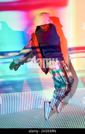 Full body of confident young ethnic female millennial in fashionable outfit and hat standing on tiptoes near wall with colorful projection and looking Stock Photo