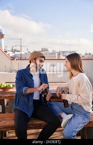 Positive young male and female flatmates sitting on bench with bottles of beer and having pleasant conversation on terrace Stock Photo