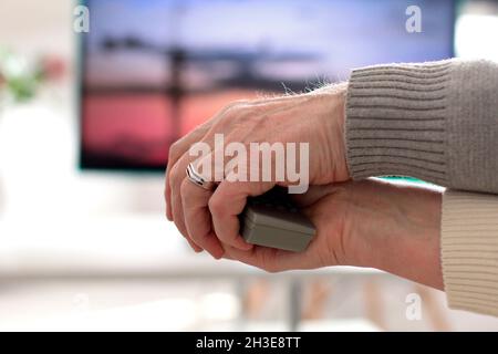 Elderly couple holding remote together with their hands while watching movie. Old man helping his wife switching channels with TV remote. || Model approval available Stock Photo