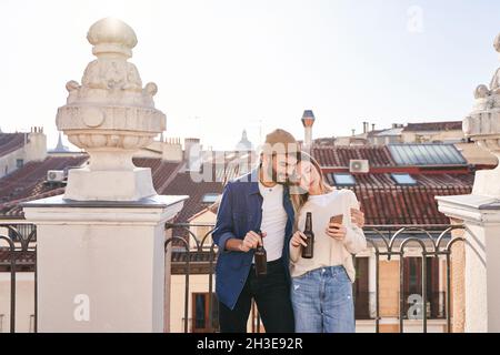 positive young friends using cellphone together while drinking beer from glass bottles and leaning on fence on balcony Stock Photo