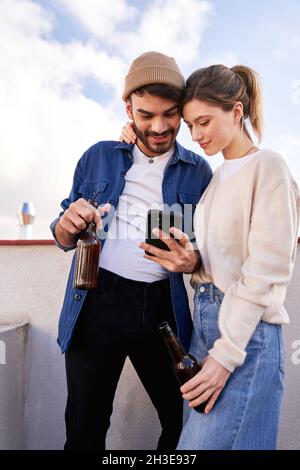 Smiling bearded man with bottle of beer hugging positive girlfriend scrolling mobile phone on balcony in sunny day Stock Photo