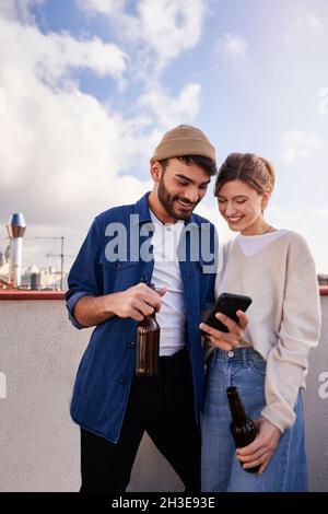 Smiling bearded man with bottle of beer hugging positive girlfriend scrolling mobile phone on balcony in sunny day Stock Photo
