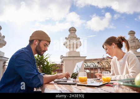 Concentrated female in eyeglasses writing in planner at table with male flatmate in wireless earbuds typing on laptop on balcony Stock Photo